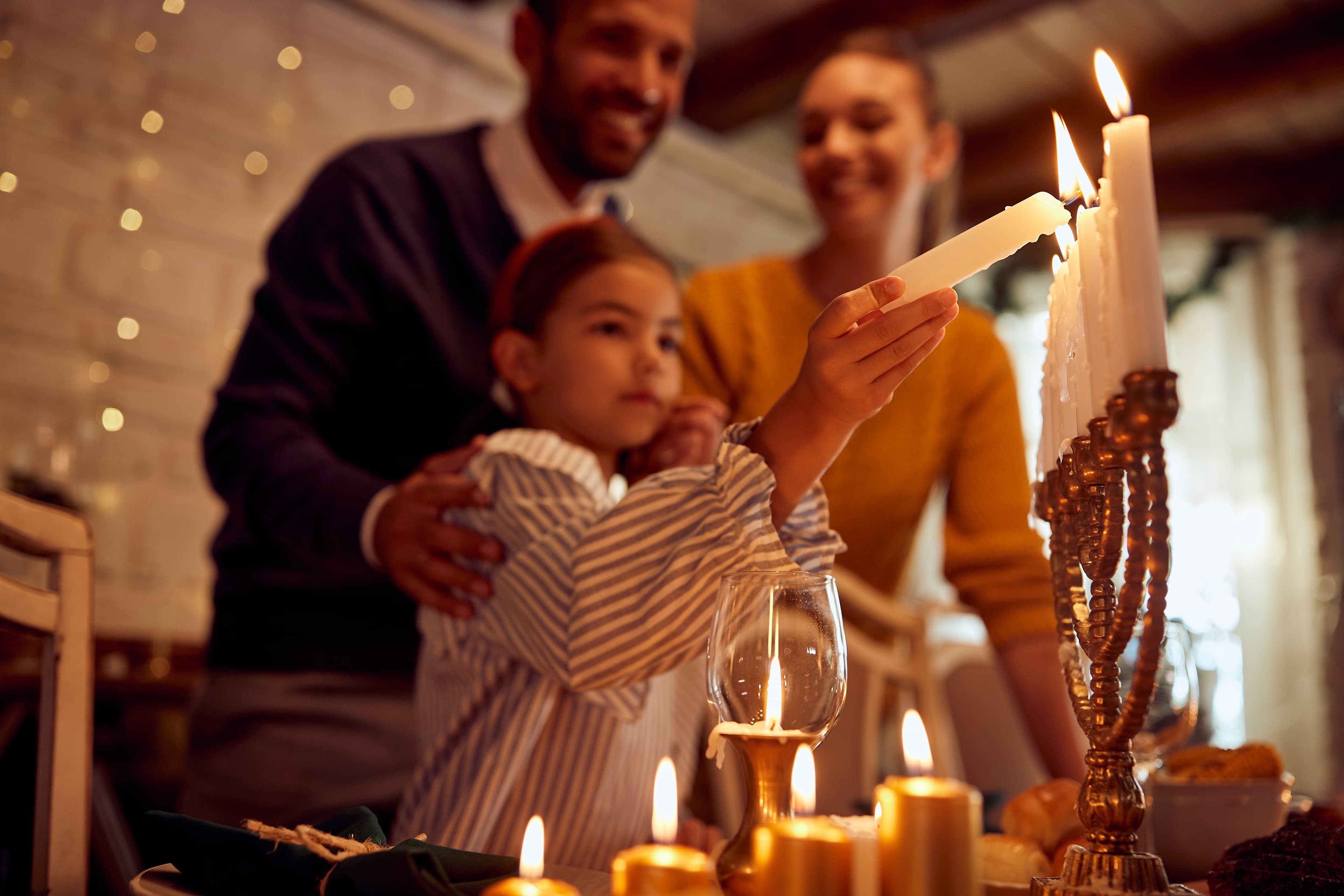 A family celebrating Hanukkah, with a young girl lighting a candle on a Hanukkiah while being supported by her parents.